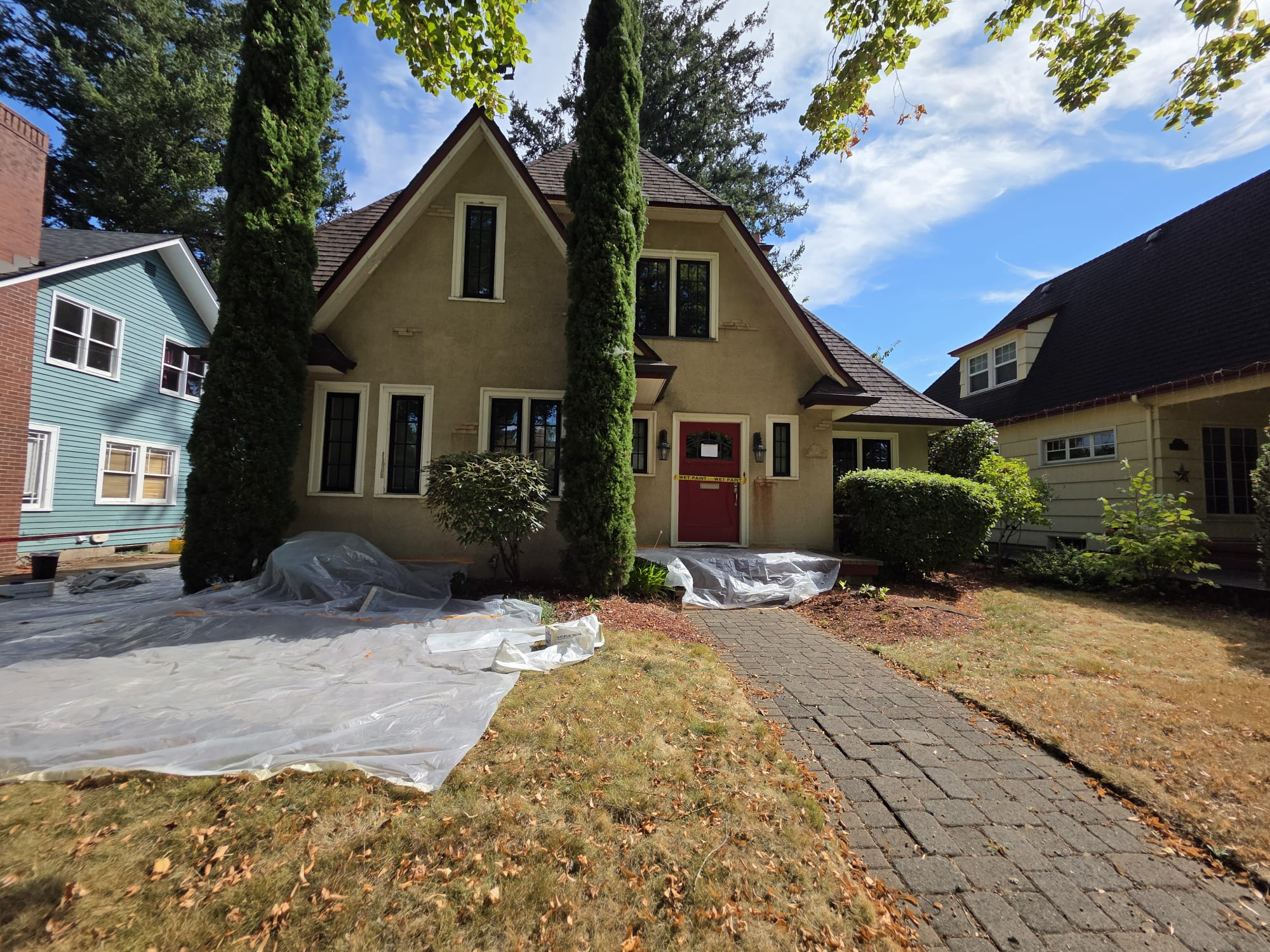 Before: two-story beige home with a red front door, protective sheeting laid over the ground and landscaping during prep.